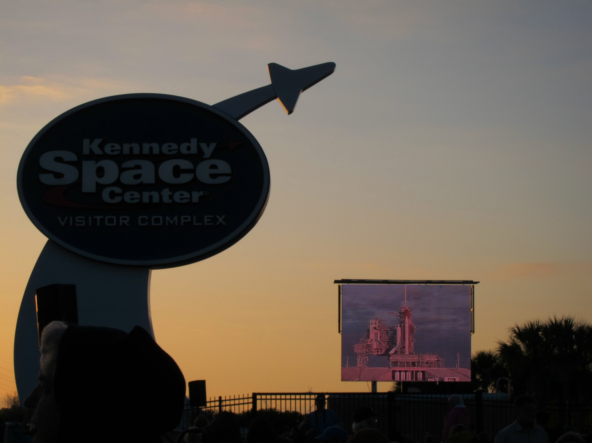 Kennedy Space Center sign silhouetted at sunset with a large screen showing the shuttle on the launch pad